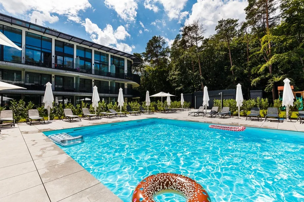 Basen hotelowy z czystą błękitną wodą otoczony leżakami i parasolami, z nowoczesnym budynkiem hotelowym w tle i bujną zielenią drzew, na pierwszym planie kolorowy dmuchany pierścień do pływania