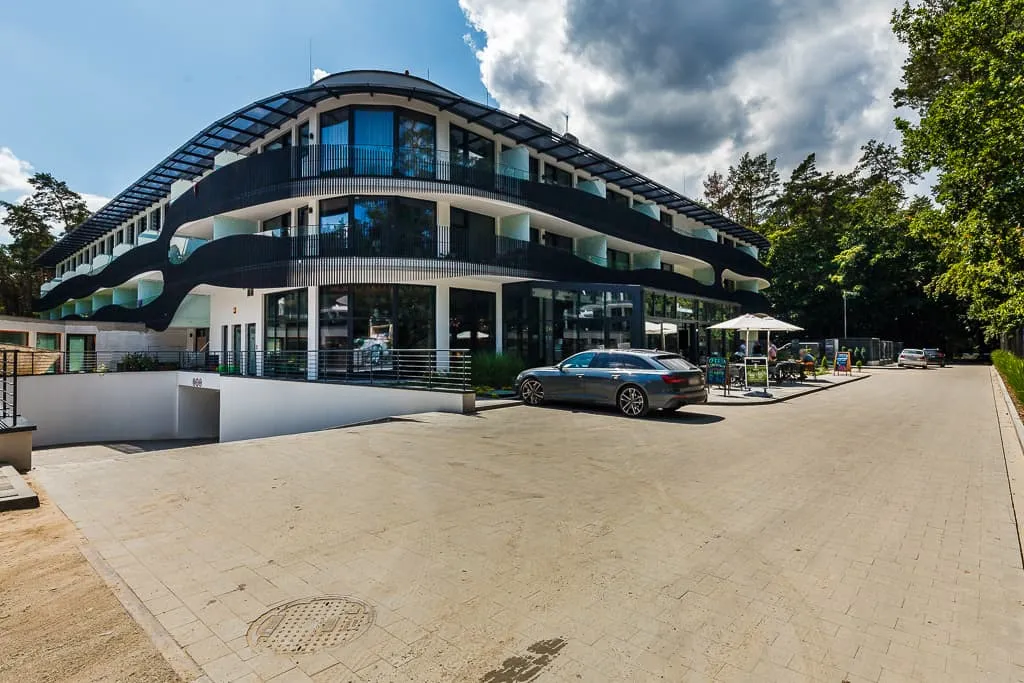 Nowoczesny budynek apartamentowy w Stegnie z białą elewacją, falistymi balkonami i dużymi przeszkleniami, z eleganckim wejściem i przestrzenią zagospodarowaną zielenią, położony w otoczeniu sosnowego lasu.